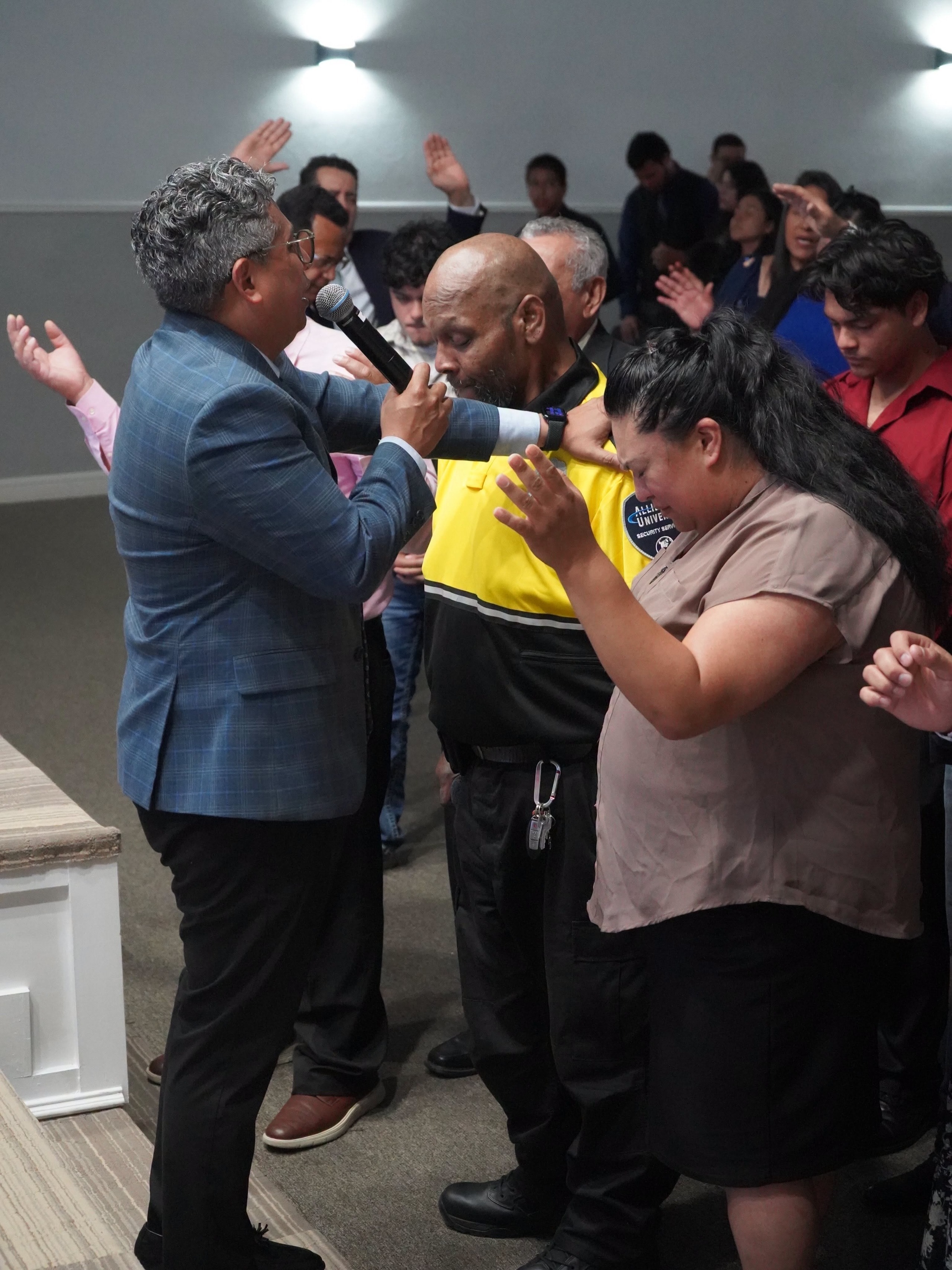 El pastor, micrófono en mano, orando por un hombre con chaleco de seguridad y una mujer mientras la congregación levanta las manos detrás de ellos